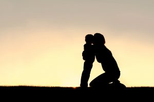 A silhouette of a young mother sitting outside, lovingly hugging her 2 year old toddler sun at sunset, with copy-space in sky.